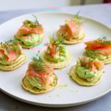 Savory quark blinis are pictured, layered with smoked salmon, avocado cream, and fresh dill.